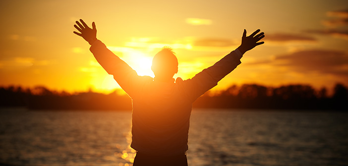 silhouette of man standing in front of water during sunset with hands stretched overhead symbolizing gratitude