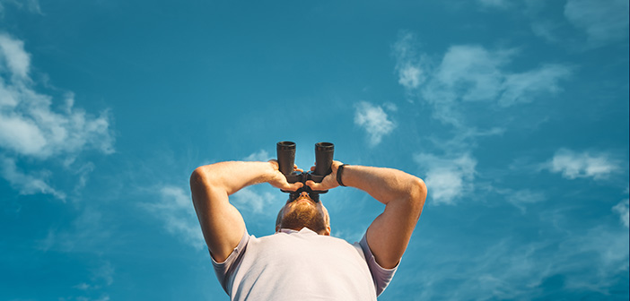 Male Traveler Looking Through Binoculars In The Distance Against The Sky
