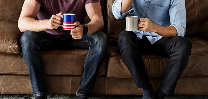 two men wearing casual clothes sitting on a brown couch drinking coffee from mugs and talking